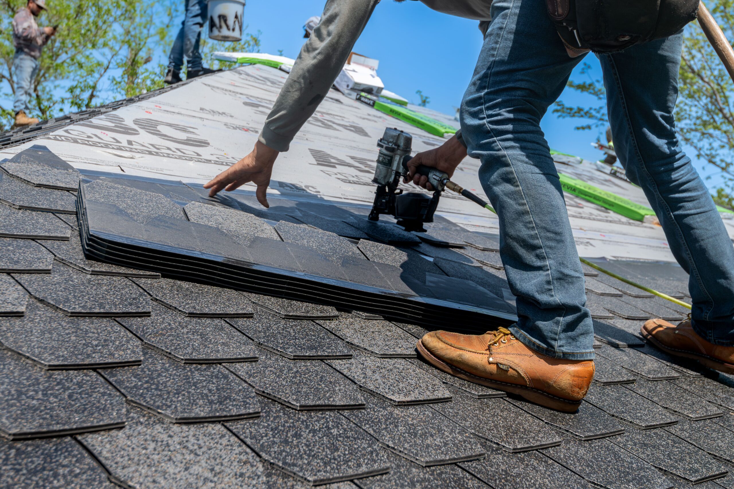 pexels-photo-33404248 - Scherger Roofing - Scherger Roofing Roofer on a pitched roof placing asphalt shingles with a nail gun nearby and bundles of shingles on the roof under a clear blue sky. | scherger roofing | scherger roofing