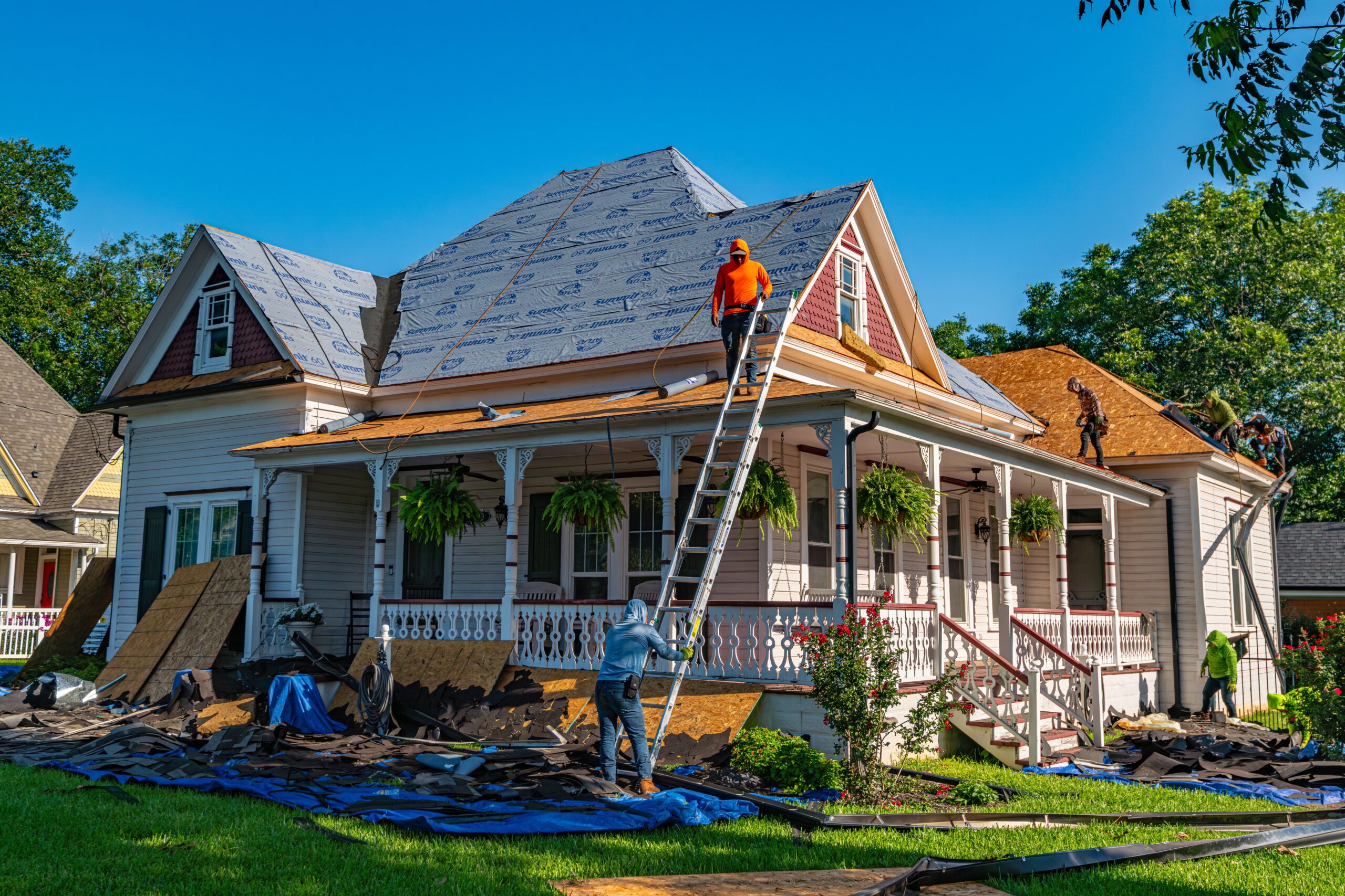 Worker on a ladder replacing shingles on a suburban house roof during a renovation, with debris scattered on the yard below under a clear blue sky | scherger roofing