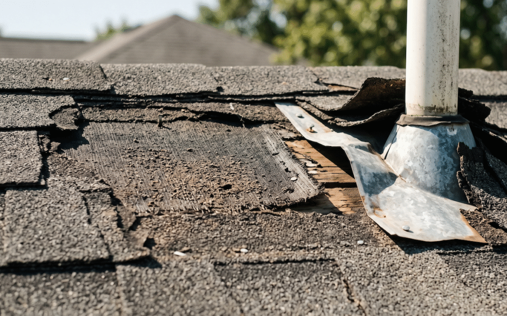 Damaged roof with peeling asphalt shingles and exposed underlayment around a metal vent pipe.