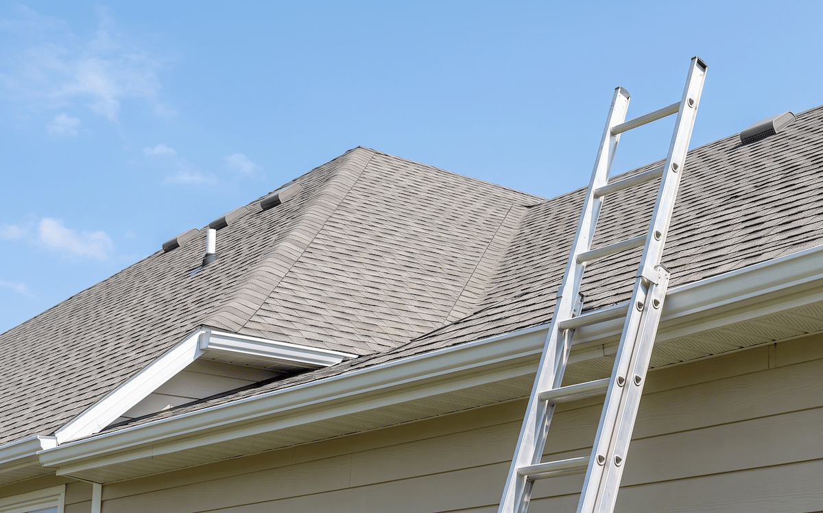Ladder leaning against a tan, pitched roof of a house under a clear blue sky, with shingles visible and gutters along the edge.