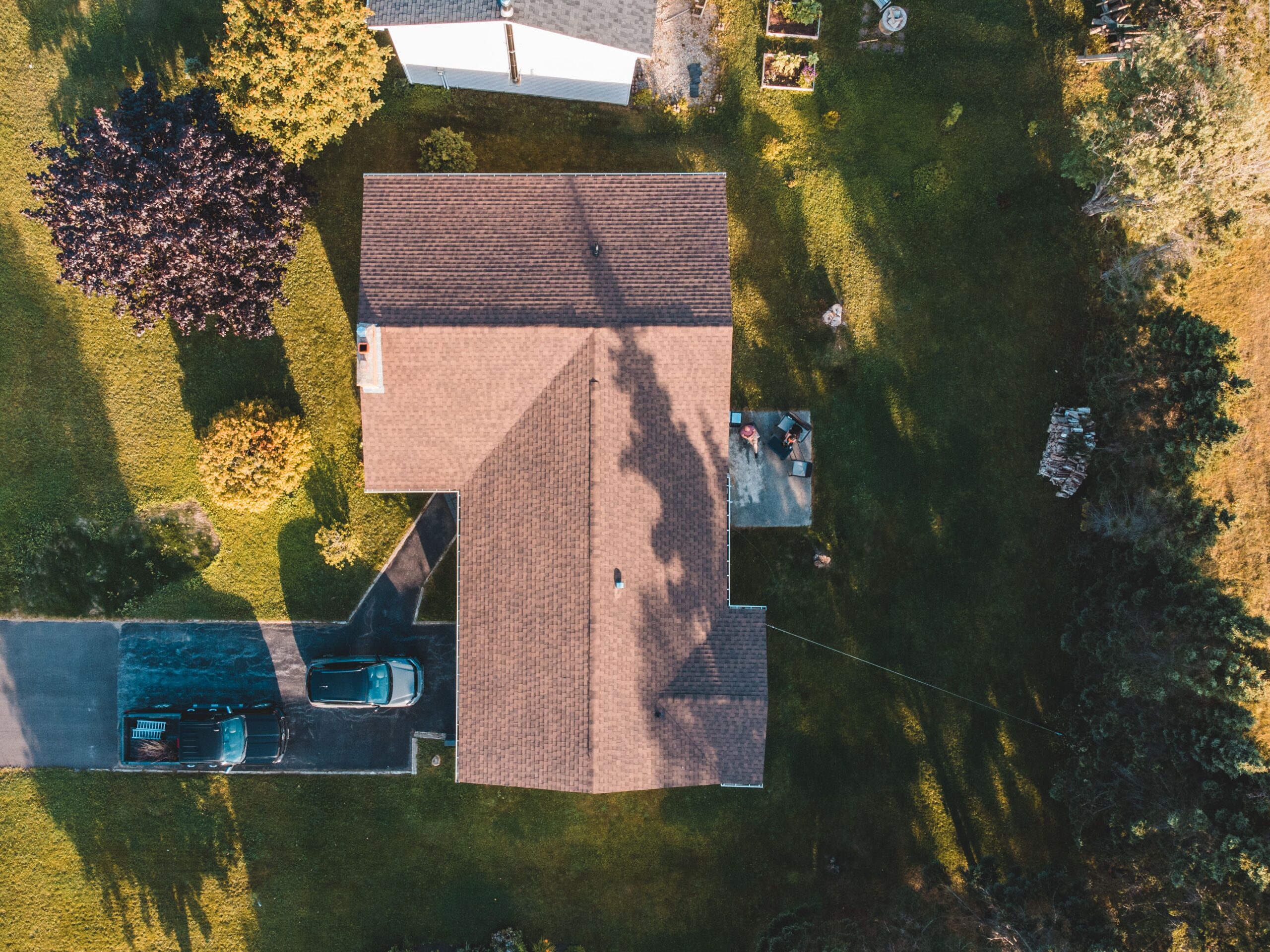Aerial view of a house with a brown shingled roof, surrounded by green lawn and trees, with a driveway and parked cars at the bottom left. | scherger roofing